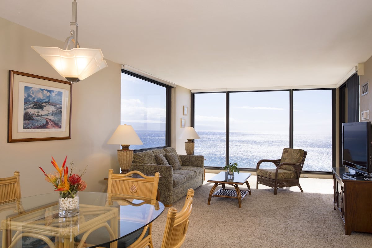 A spacious living room with a large window overlooking the ocean, a comfortable sofa, a dining table with chairs, and a television on a wooden stand.