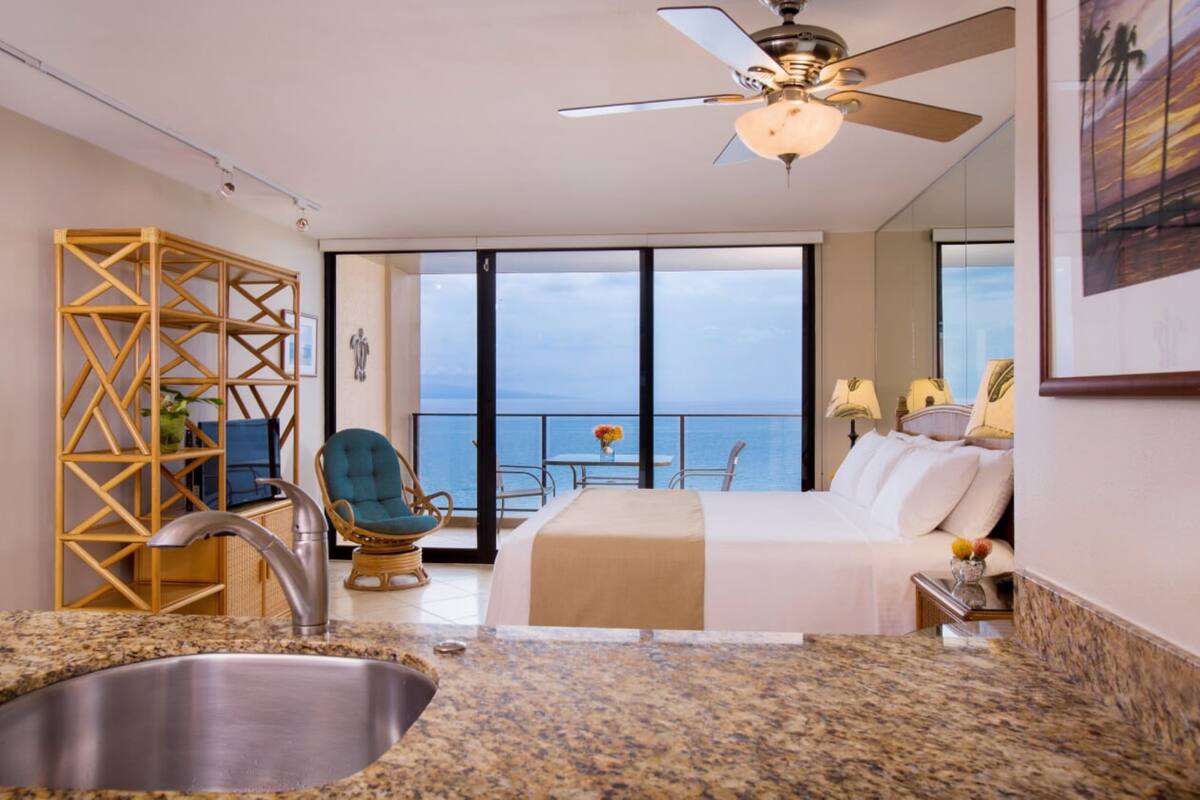 A hotel room with a bed, a sink, a balcony with a view of the ocean, and a ceiling fan.