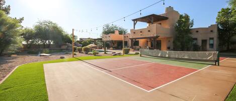 An outdoor tennis court with a net, surrounded by a landscaped area with trees and a building in the background.