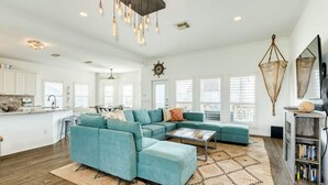 A spacious living room with a teal sectional sofa, a coffee table, and a patterned rug. There is a kitchen area with white cabinets and a large window. - Captain's Quarters - Cabana, Hot Tub, Fire Pit, Fenced Yard! (Freeport)
