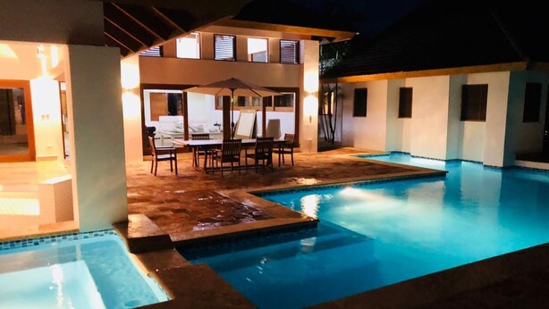 Ein Schwimmbecken mit einer Terrassenanlage, einer überdachten Lounge-Bereich und einem gut beleuchteten Gebäude im Hintergrund.