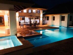 Une piscine avec un espace patio, un coin salon couvert et un bâtiment bien éclairé en arrière-plan.