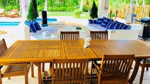 Ein überdachter Außenbereich mit Essbereich, Holztischen und -stühlen, einem Pool und einer Lounge mit blauen Kissen.