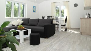 A modern living room with a grey sofa, a small white coffee table, and a black ottoman. There is a dining area with a white table and chairs, and a kitchen area with a wooden cabinet and a clock on the wall. - Mocici apartments (Konavle)