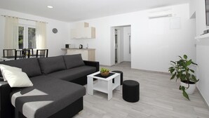 A modern living room with a grey sectional sofa, a small white coffee table, and a black ottoman. There is a dining area with a table and chairs, and a kitchen area with cabinets and a sink. - Mocici apartments (Konavle)