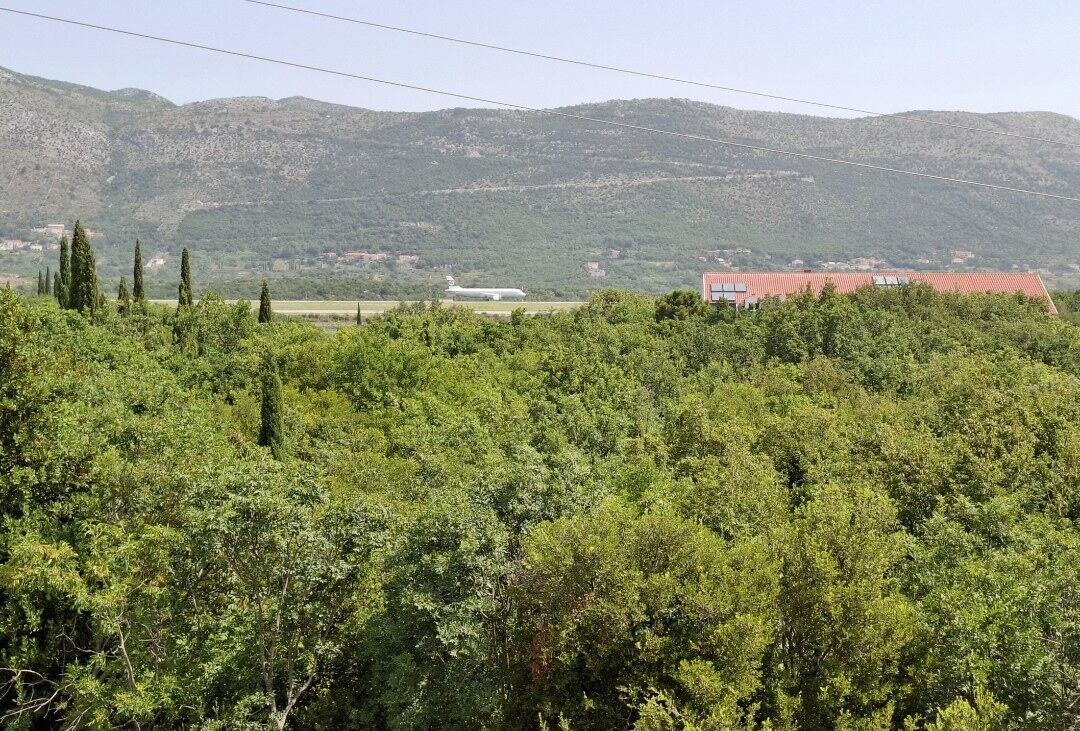 Paesaggio rurale con un edificio a tetto rosso, circondato da vegetazione lussureggiante e colline sullo sfondo.
