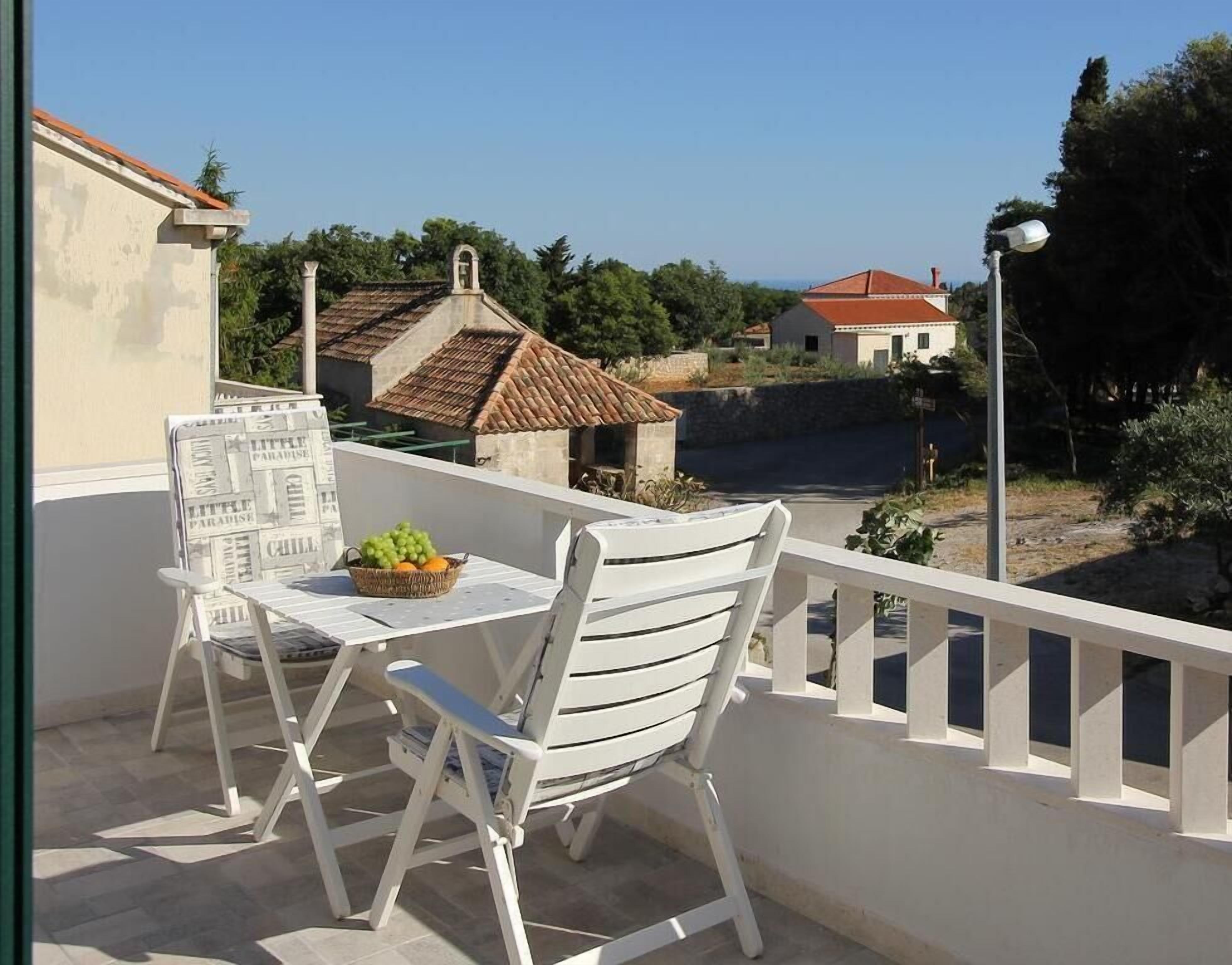 Un balcón con muebles blancos, una mesa con una cesta de fruta, y vistas a casas y árboles.
