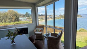 Ein Zimmer mit großem Fenster, das auf ein Gewässer und eine Wiese blickt. Darin stehen Tisch und Stühle sowie ein Blumenstrauß.