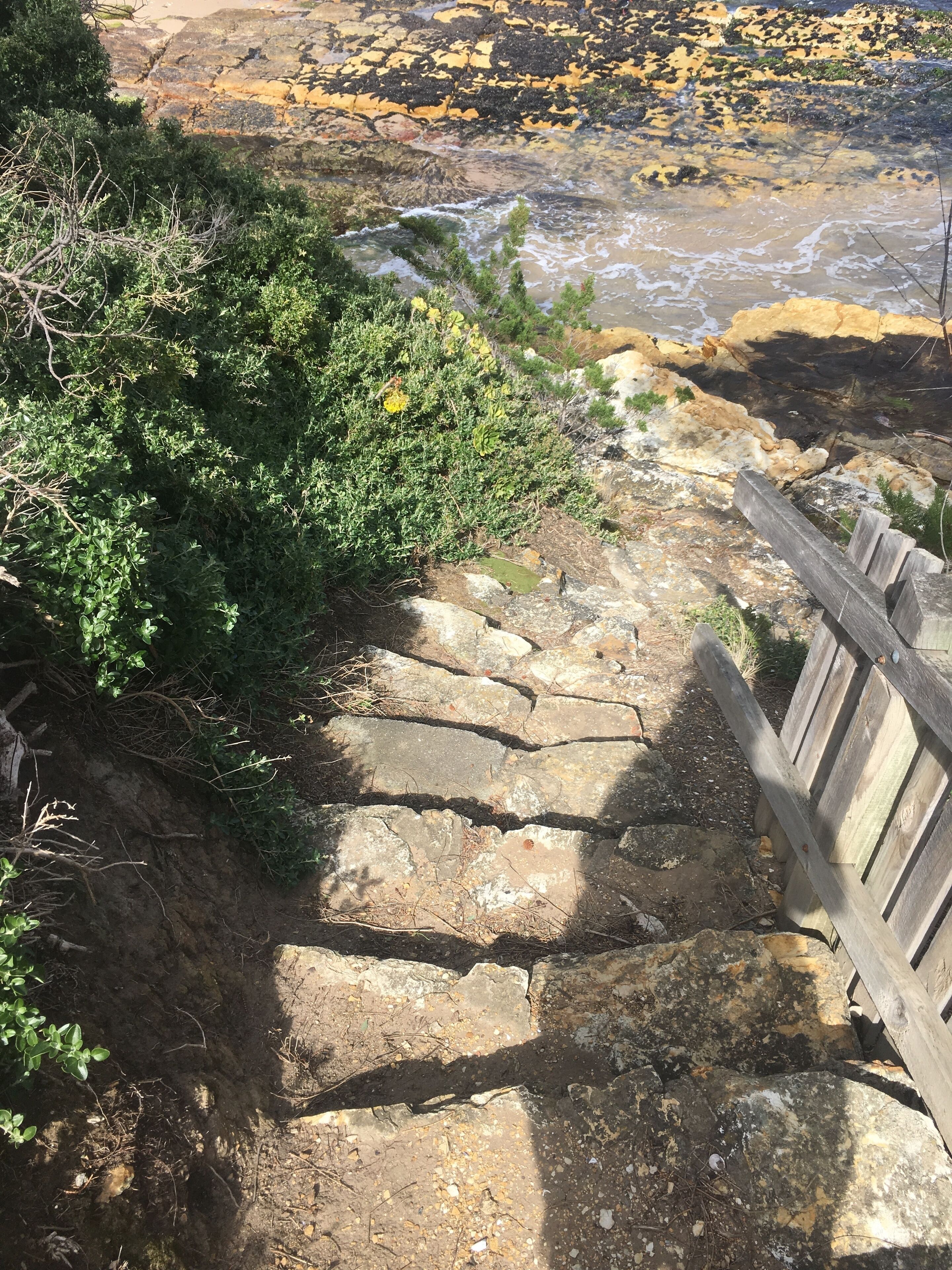 Eine steile Steintreppe führt zu einer felsigen Küstenlinie, auf der rechten Seite verläuft ein Holzgeländer.