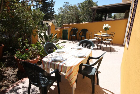 An outdoor patio with a table set for four, surrounded by chairs, and a potted plant to the left.