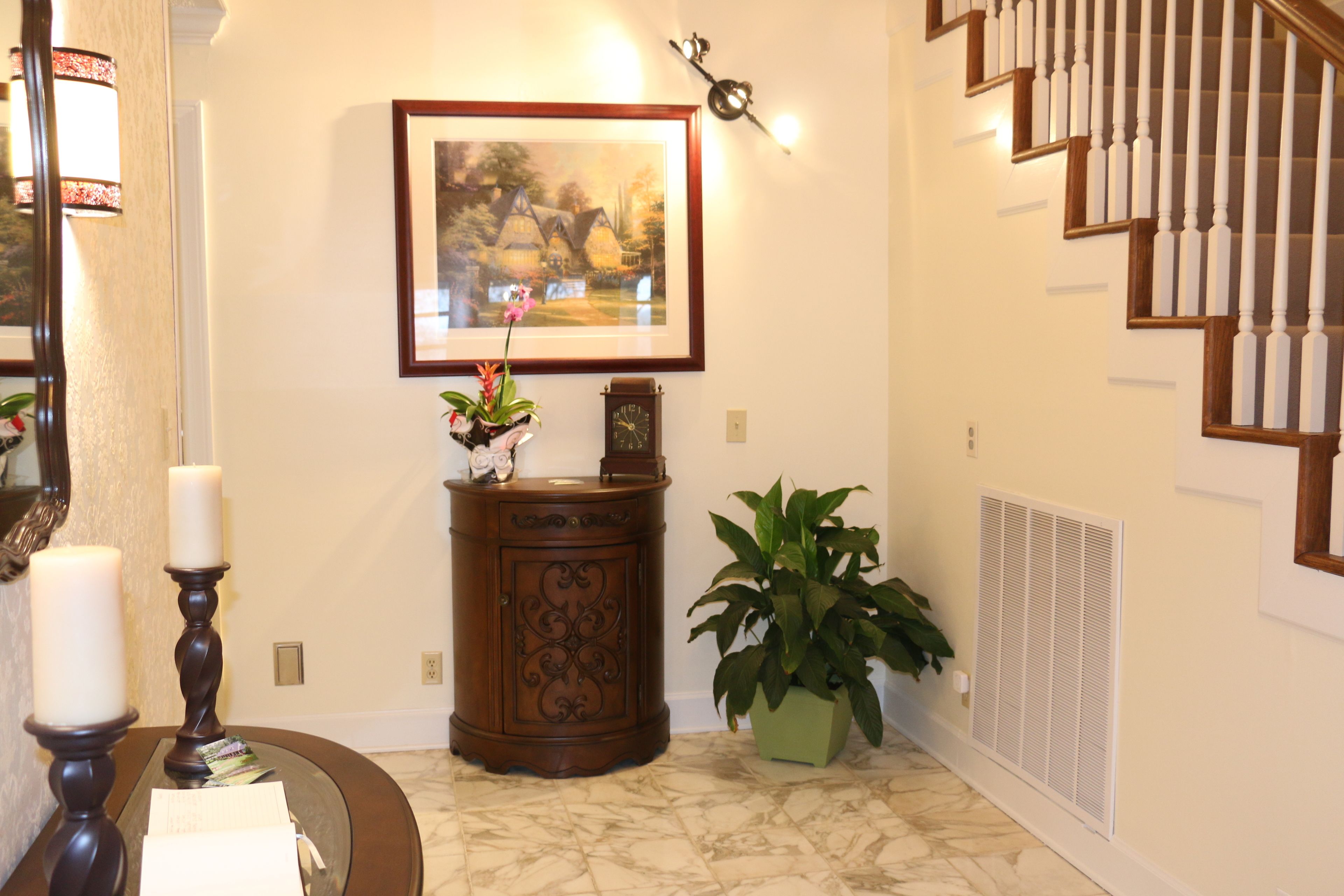 A hallway with a framed painting, a wooden cabinet, a potted plant, a mirror, and a staircase.