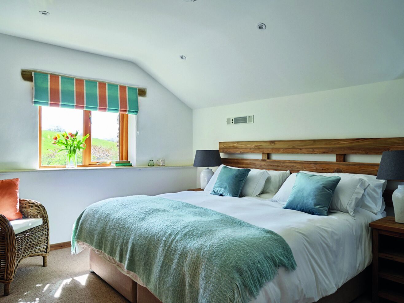 A bedroom with a large bed, a window with a view of greenery, a wicker chair, and a bedside table with a lamp.