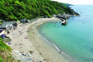 A coastal beach with clear turquoise waters, a curved shoreline, and a stone wall separating the beach from the forested hillside. - Stunning Barn Conversion, Indoor Pool, tranquil vineyard site, coastal location. (Fowey)