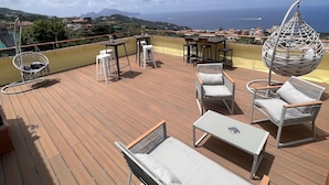 A wooden deck with outdoor furniture, including lounge chairs and a hanging chair, overlooking a coastal view with mountains and the sea. - Relais il Chiostro Historic Courtyard and Panoramic Terrace near Sorrento (Massa Lubrense)