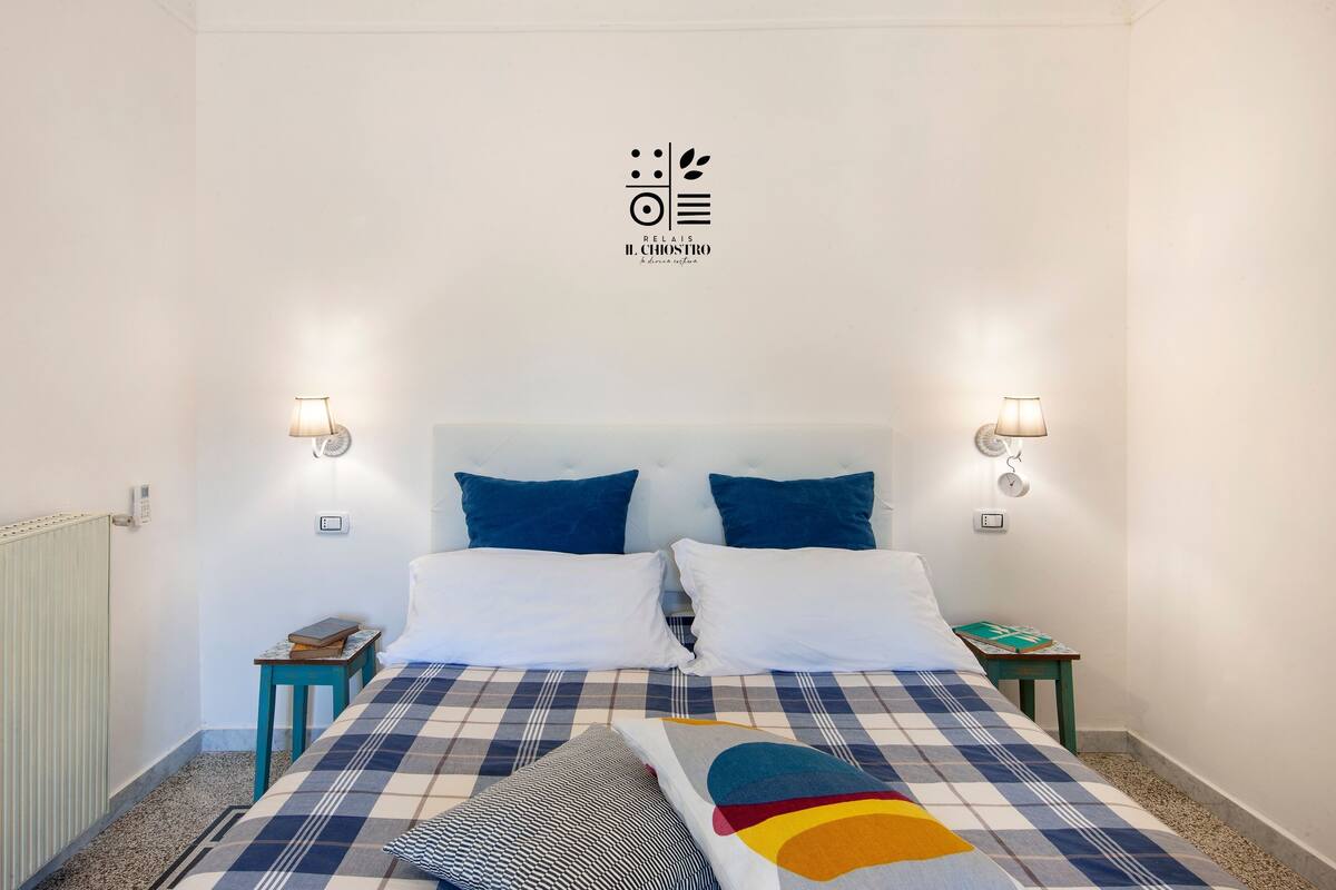 A neatly arranged bedroom with a checkered bedspread, two pillows, a bedside table with a book, and wall-mounted lamps.
