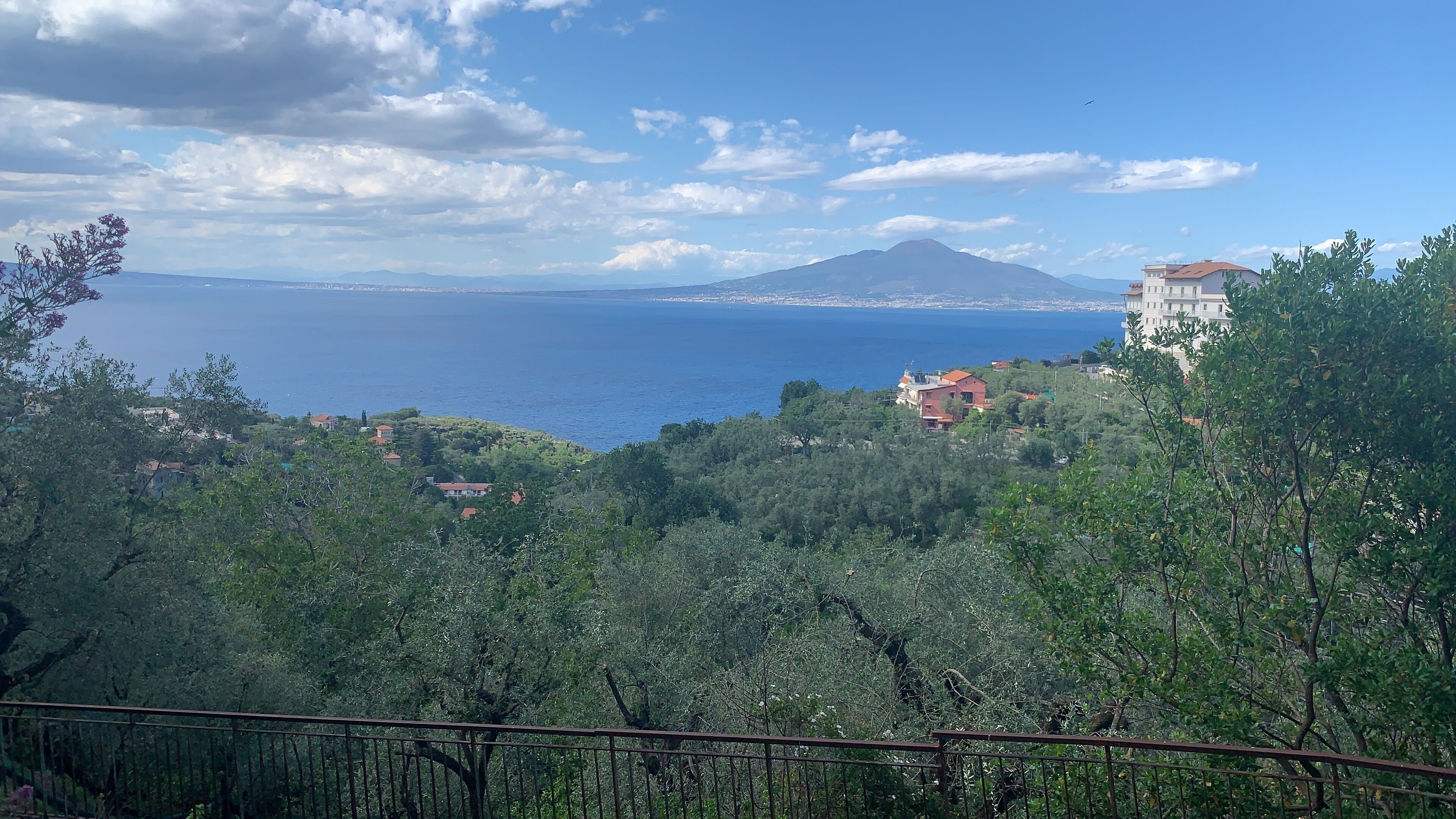 A coastal landscape with a mountain in the background, houses on a hillside, and a clear view of the sea.