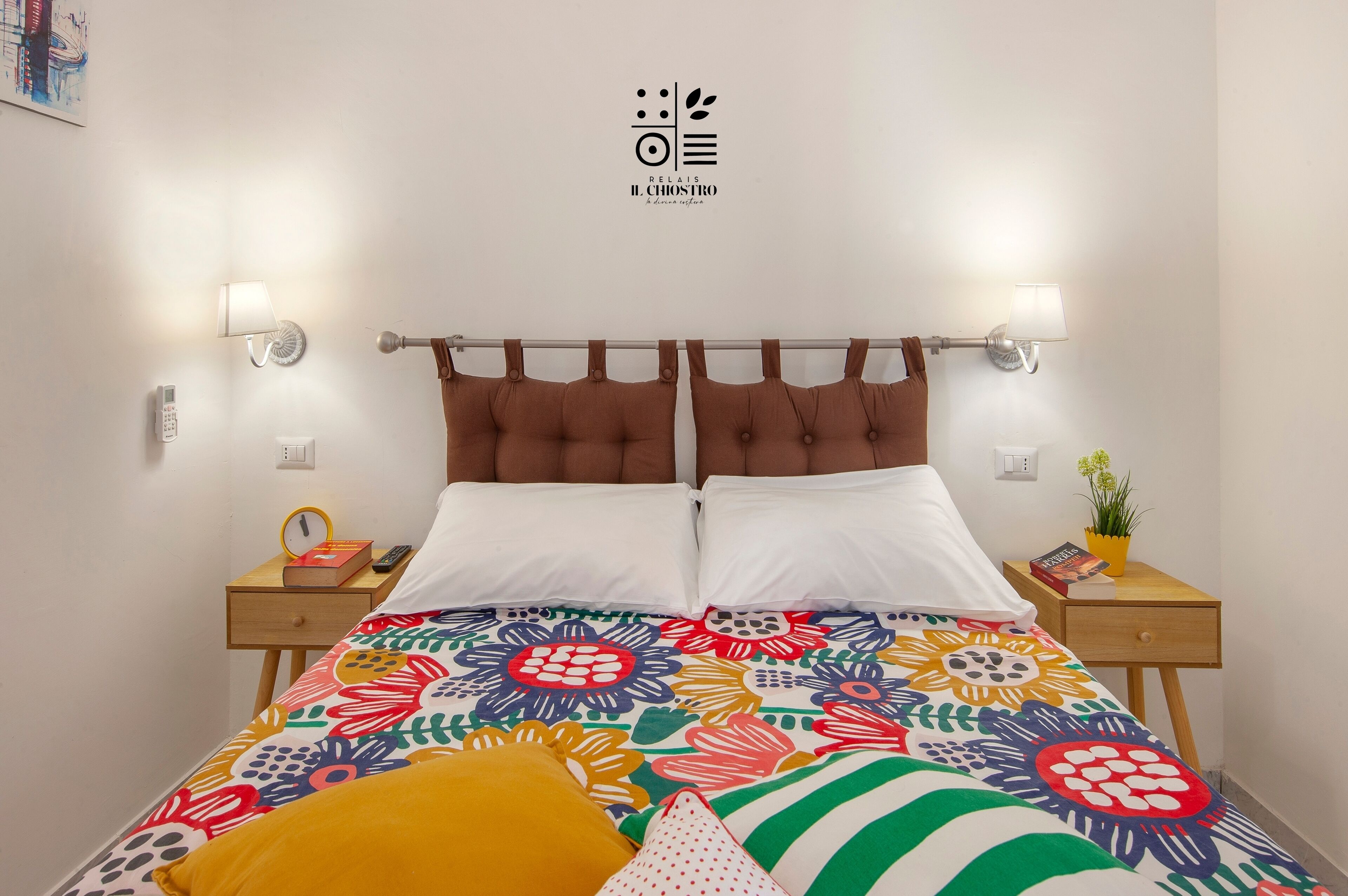 A neatly made bed with a colorful floral bedspread, two bedside lamps, a small bedside table with a book and a plant, and a wall-mounted logo.