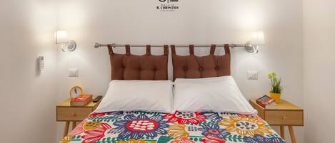 A neatly made bed with a colorful floral bedspread, two bedside lamps, a small bedside table with a book and a plant, and a wall-mounted logo.