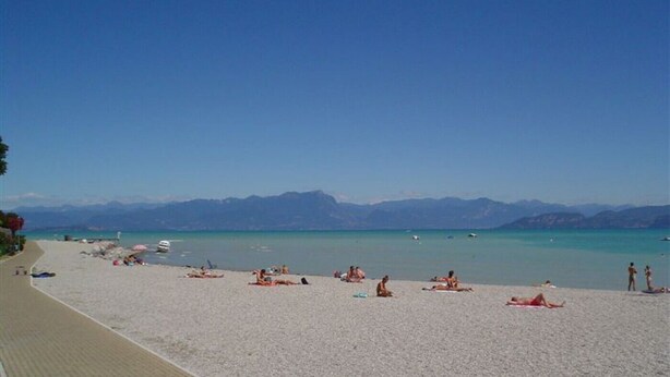 Een strand met mensen die zonnebaden en zwemmen, een heldere lucht en bergen op de achtergrond.