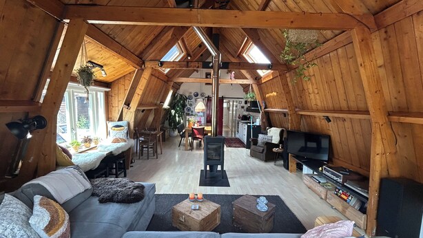 En stue i skråvæg med sofa, sofabord og fjernsyn. Der er et spiseområde med bord og stole, og et køkken i baggrunden.