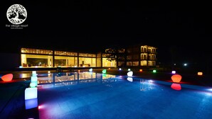 A nighttime view of a resort with a swimming pool, illuminated by colorful lights, and a building with traditional architecture. - The Village Resort (Ngwe Saung)