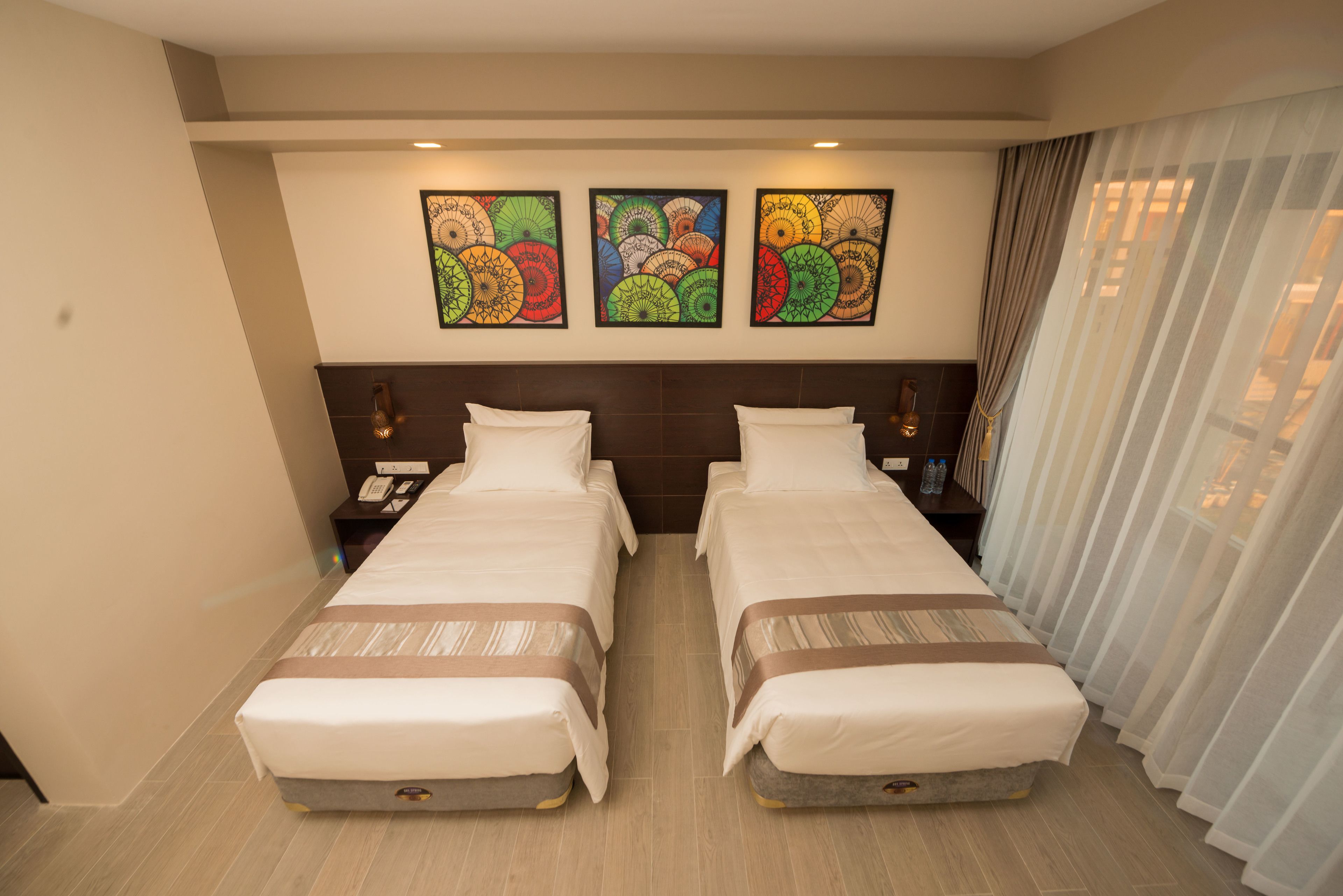 A hotel room with two beds, a headboard with artwork, a bedside table with a lamp, and a window with curtains.