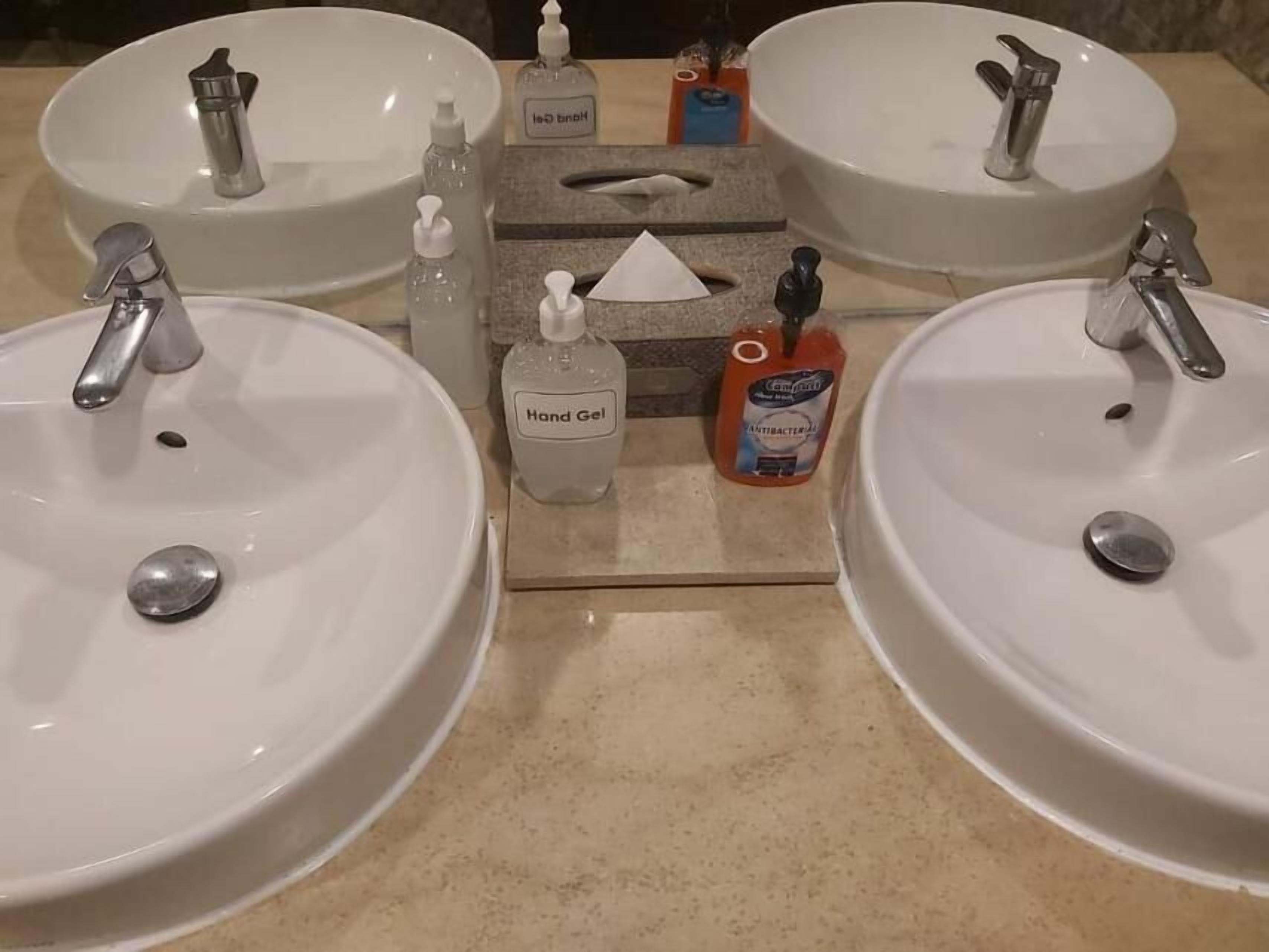 A bathroom with two sinks, each with a faucet and a soap dispenser. There is a hand gel dispenser and an antibacterial soap bottle on the counter.