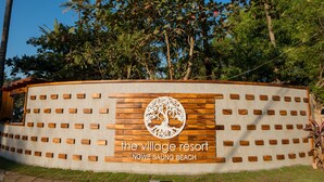 A sign for The Village Resort at Ngwe Saung Beach, featuring a wooden panel design with a tree logo. - The Village Resort (Ngwe Saung)
