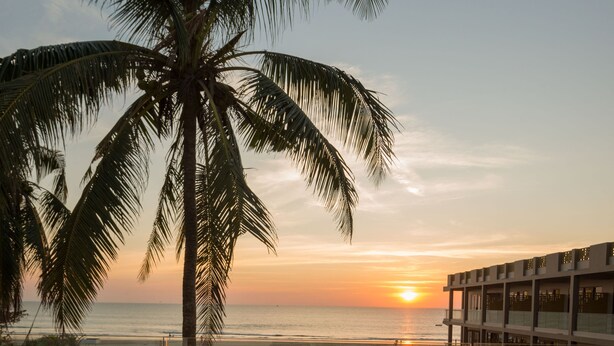 A balcony with a wooden table and two chairs, overlooking a beach and palm tree at sunset. - The Village Resort (Ngwe Saung)