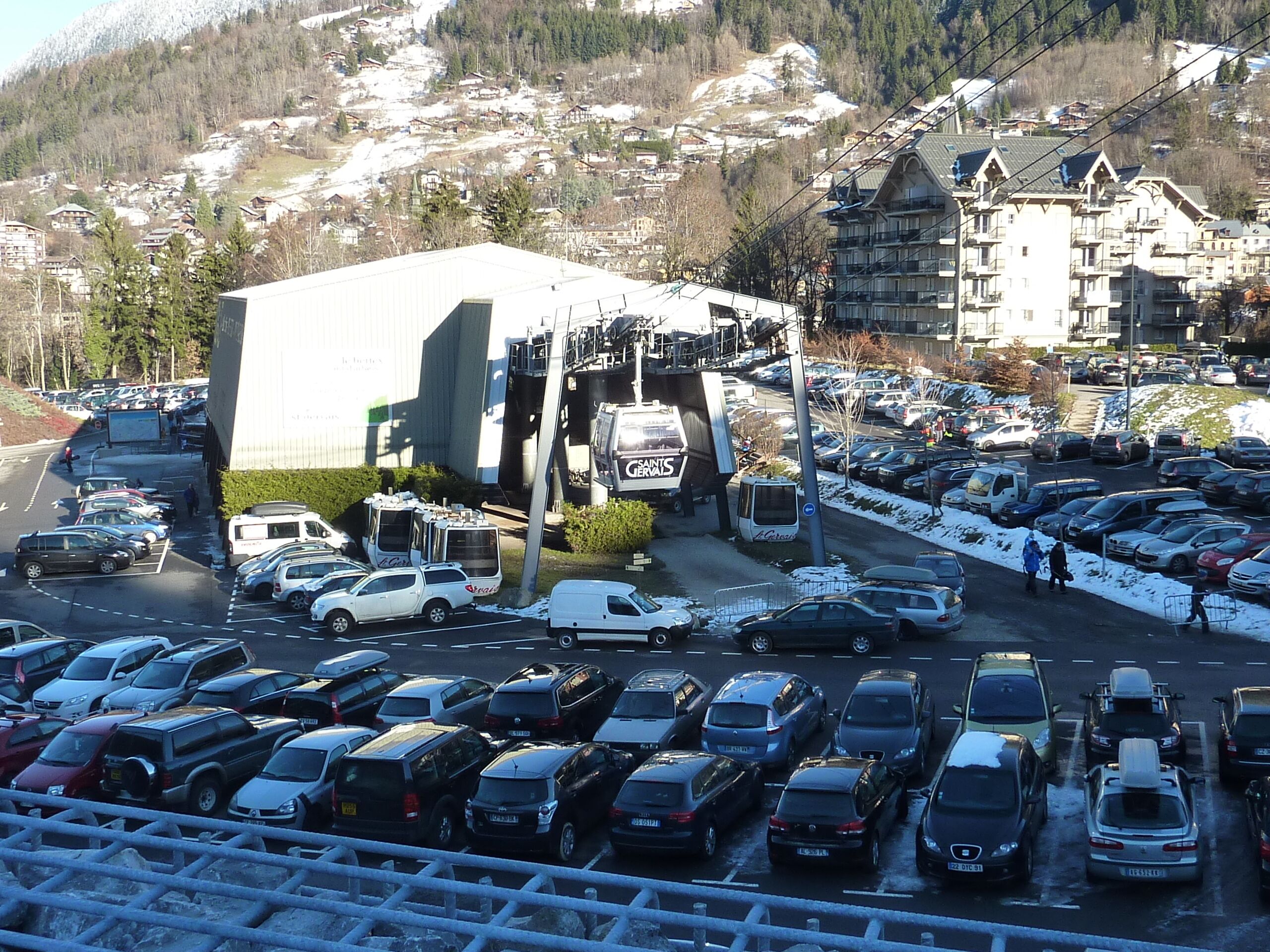 Un parcheggio con diverse auto e un autobus, circondato da edifici e montagne sullo sfondo.