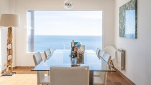 A dining area with a glass table, white chairs, a view of the sea, a lamp, and a radiator.