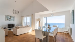 A modern living room with a TV, sofa, and dining area with a view of the sea.