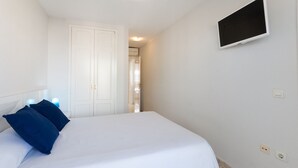 A bedroom with a bed, two blue pillows, a white headboard, a white door, a TV mounted on the wall, and a light switch. - Beautiful property with 2 bedrooms. Swimming pool. Wifi - only families (Playa de Gandía)