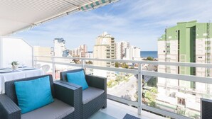 A balcony with wicker furniture and a table set for two, overlooking a cityscape with buildings and a view of the sea. - Beautiful property with 2 bedrooms. Swimming pool. Wifi - only families (Playa de Gandía)
