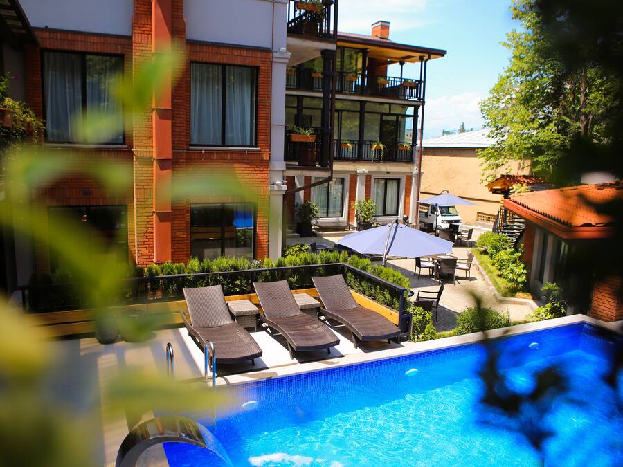A rooftop pool with lounge chairs, a dining area, and a view of a multi-story building with balconies.