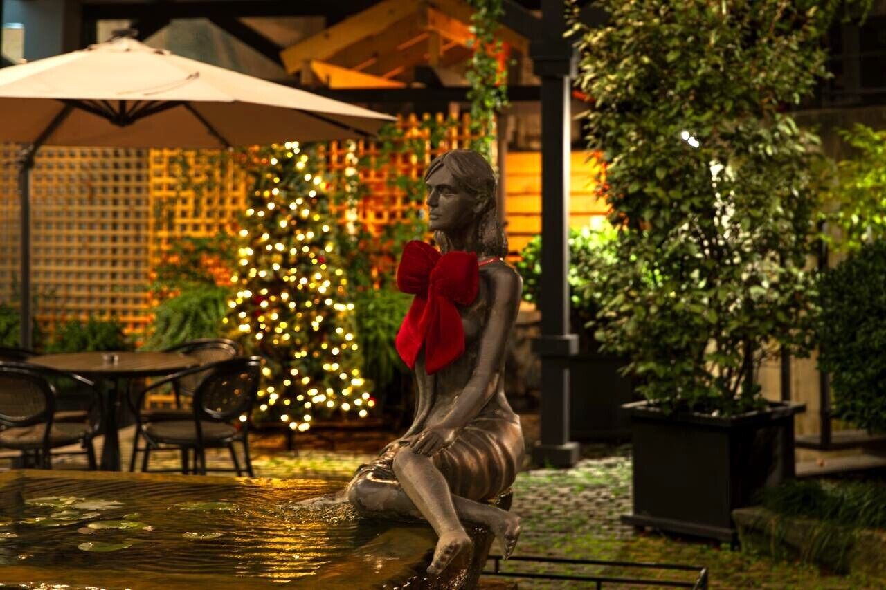 Eine Statue einer Frau in einer Fontäne, geschmückt mit einer roten Schleife, umgeben von Außenplätzen und einem Weihnachtsbaum.