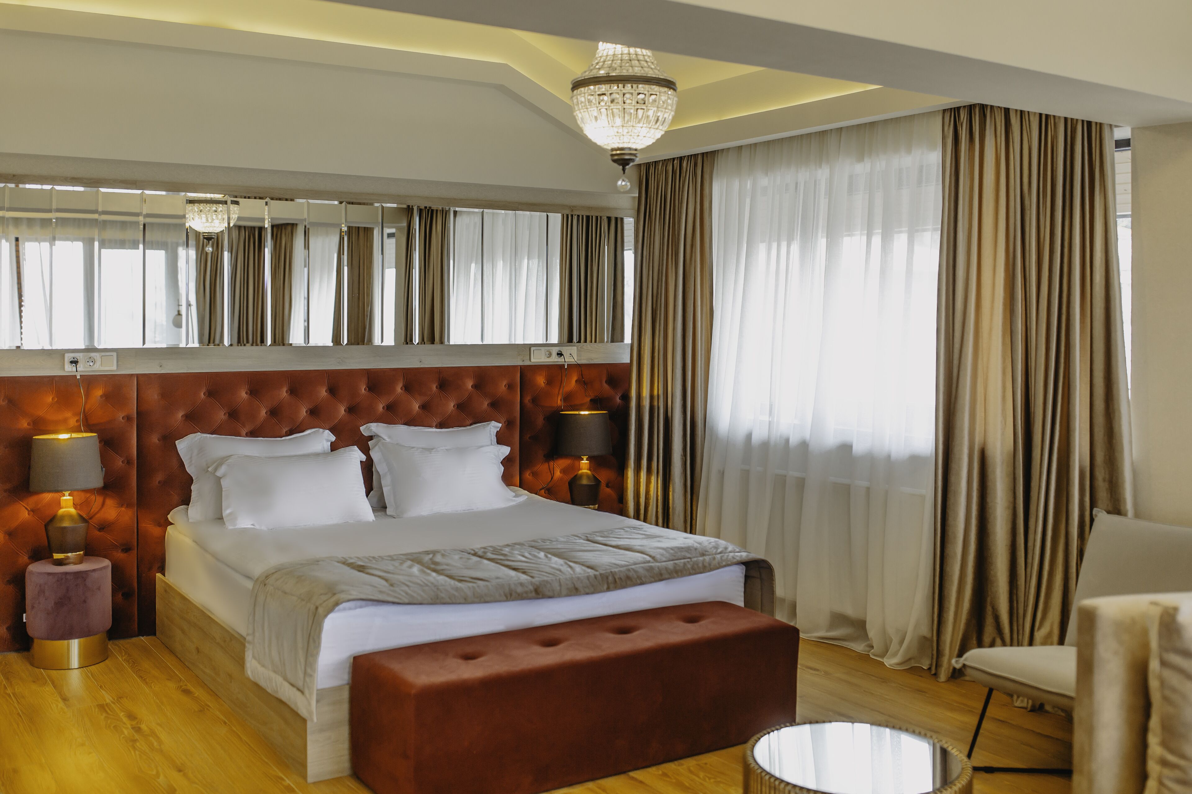 Een slaapkamer met een gestoffeerd hoofdeinde, een groot bed, twee bedlampen, een ronde spiegel, een houten vloer en een groot raam met gordijnen.