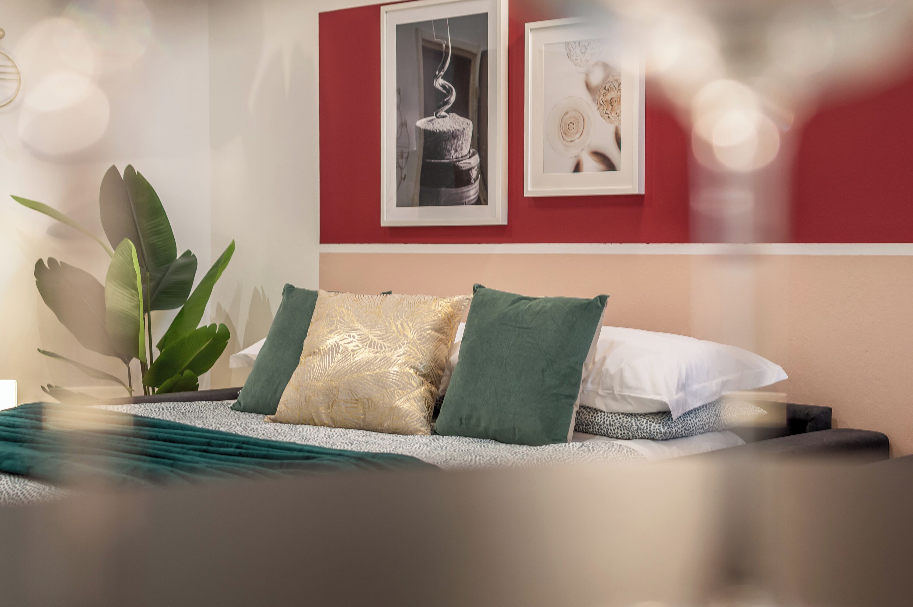 A neatly made bed with green and white pillows, a large green plant, and two framed pictures on the wall.