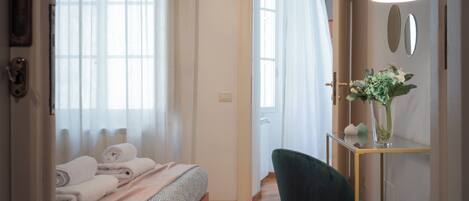A hotel room with a bed, a desk with a vase of flowers, and a door leading to another room.
