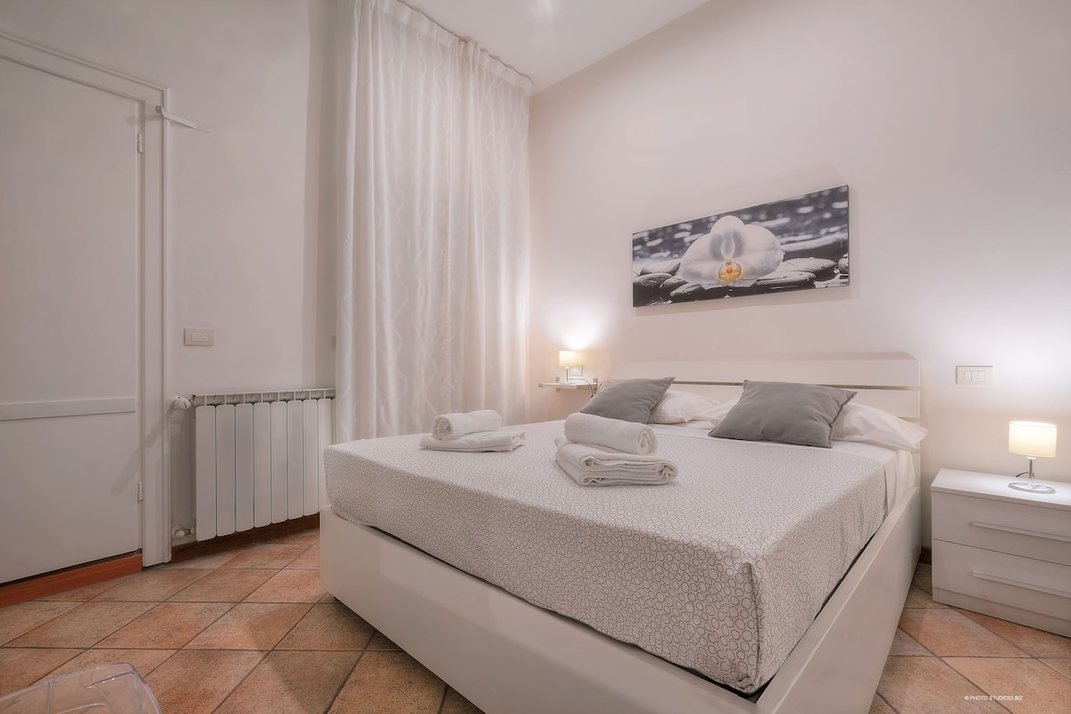 A neatly made bed with towels, a bedside lamp, and a painting of a flower on the wall.