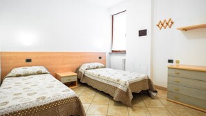 Une chambre avec deux lits simples, une tête de lit en bois, une petite table de chevet, une commode et une fenêtre donnant sur l’extérieur.