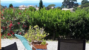 An outdoor seating area with a table and chairs, surrounded by lush greenery and a view of the sea and distant hills. - Villa with a stunning ocean view and a quiet, shaded terrace; air-conditioned bedrooms (La Croix-Valmer)