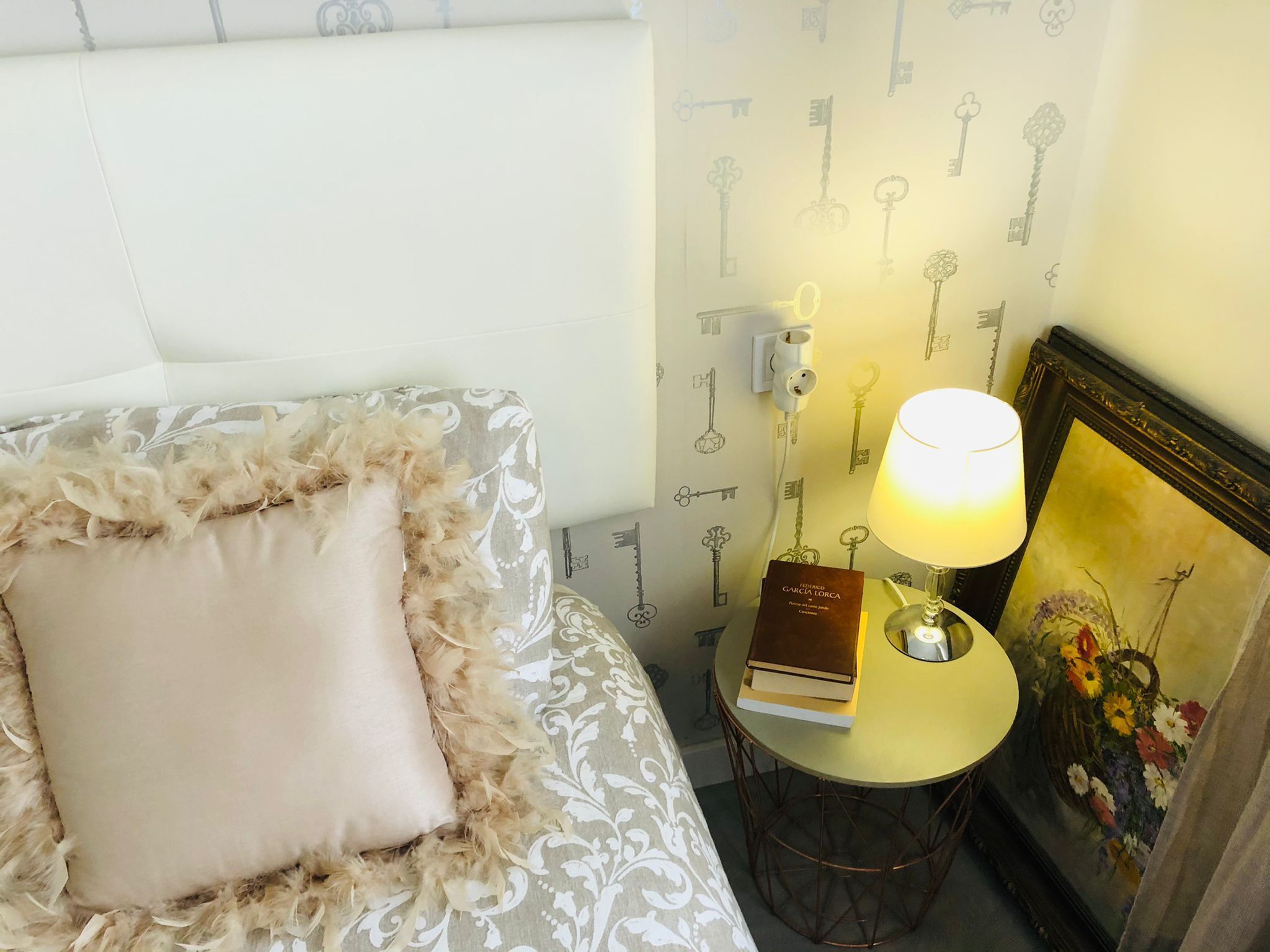Una cama bien hecha con un almohadón mullido y una colcha estampada. Junto a ella, una pequeña mesa redonda con una lámpara y un libro. La pared presenta un papel tapiz con motivos de llaves.