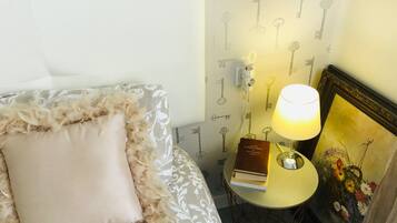 Una cama bien hecha con un almohadón mullido y una colcha estampada. Junto a ella, una pequeña mesa redonda con una lámpara y un libro. La pared presenta un papel tapiz con motivos de llaves.