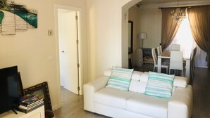Un salon avec un canapé blanc, un téléviseur et une bibliothèque. On aperçoit à l’arrière-plan un coin repas avec une table et des chaises.