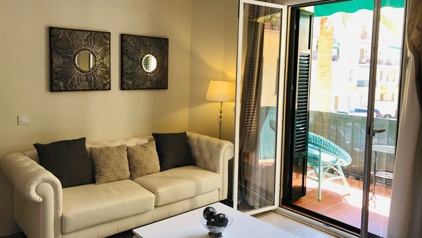 Un salon avec un canapé beige, deux miroirs muraux, une table basse blanche et une porte-fenêtre coulissante donnant sur un espace extérieur équipé d’une chaise et d’une table.