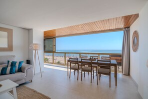 A modern living room with a sofa, a floor lamp, and a dining area with a table and chairs. A large window offers a view of the beach and ocean.