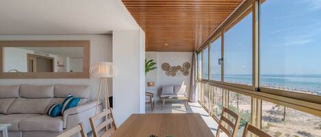 Een moderne woonkamer met een houten plafond, een groot raam met uitzicht op een strand, een eettafel met stoelen en een bank met kussens.