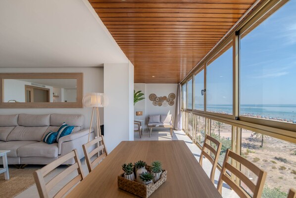 A modern living room with a wooden ceiling, a large window overlooking a beach, a dining table with chairs, and a sofa with cushions.