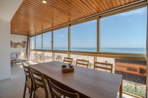 A dining area with a wooden table and chairs, large windows with a view of the beach and ocean, and a wall with a decorative pattern.